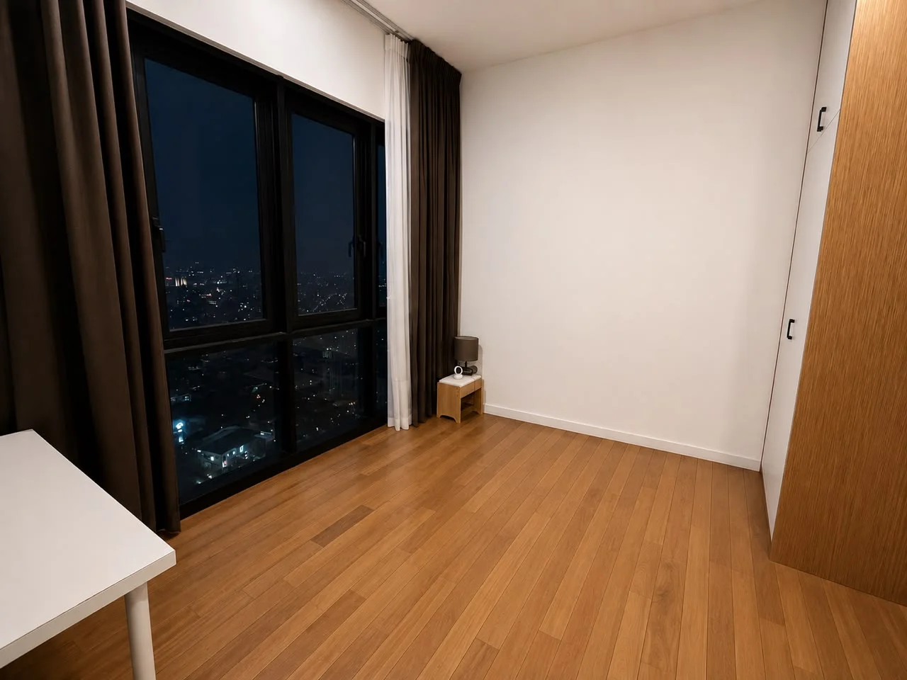 Bedroom features: wooden flooring, white painted walls, floor-to-ceiling windows, blackout curtains, night city view, built-in wardrobe, small side table, desk, minimalist decor; style: modern; view: city.